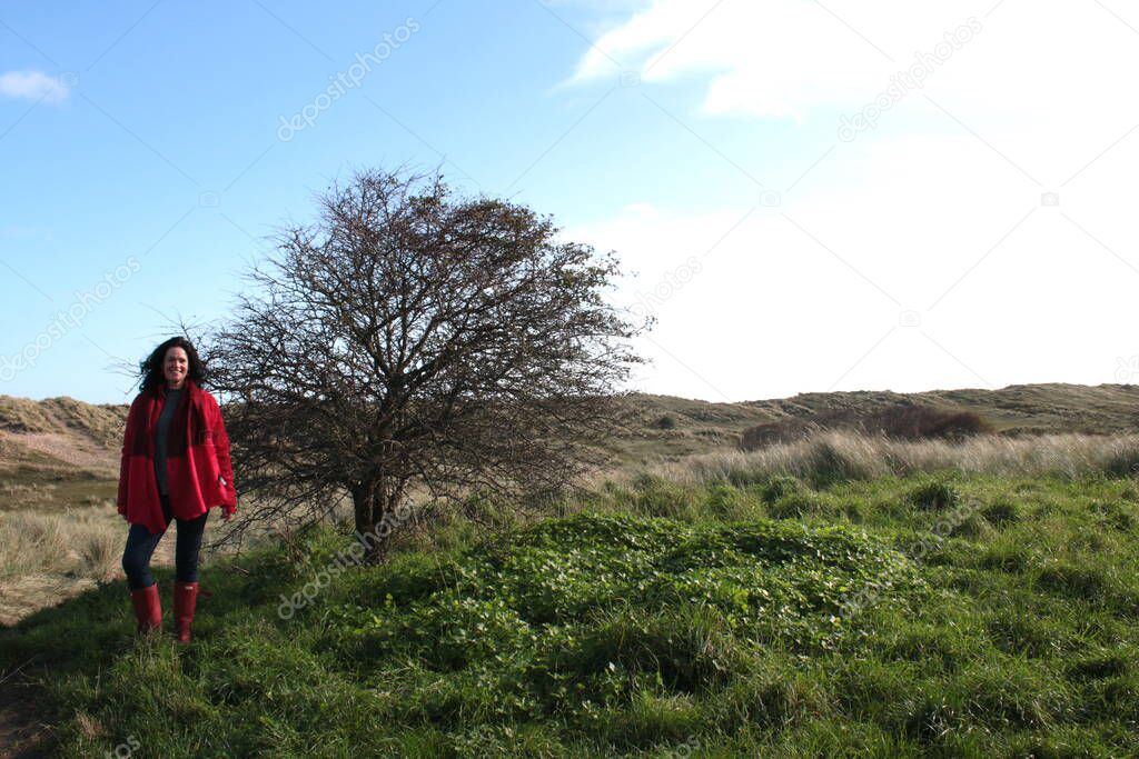 Attractive black haired female  middle aged in coast landscape standing smiling next to tree on grassy sand dunes by ocean fresh air wearing red scarf boots and coat with blue skies getting exercise during the covid-19 lockdown restrictions 2021