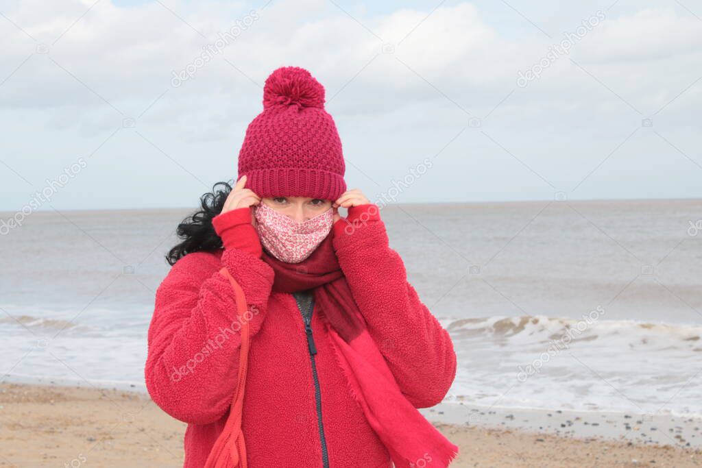 Close up of female putting on a mask with black hair in red clothes of red jacket scarf and hat seated on seaside beautiful sandy beach by ocean in Winterton Norfolk east Anglia UK  in lockdown for Covid-19 virus outbreak sea background