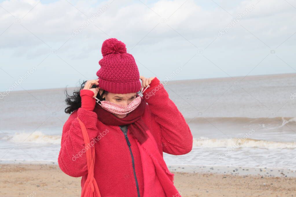 Close up of female putting on mask cover with black hair in red clothes of red jacket scarf and hat seated on seaside beautiful sandy beach by ocean sea shore in Winterton Norfolk east Anglia UK  in lockdown for Covid-19 virus outbreak sea background