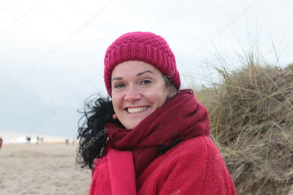 Close up portrait of attractive female smiling outdoors on beach with black hair pale skin and wearing a red scarf and jacket with sea ocean background in Autumn cold weather with sunshine 