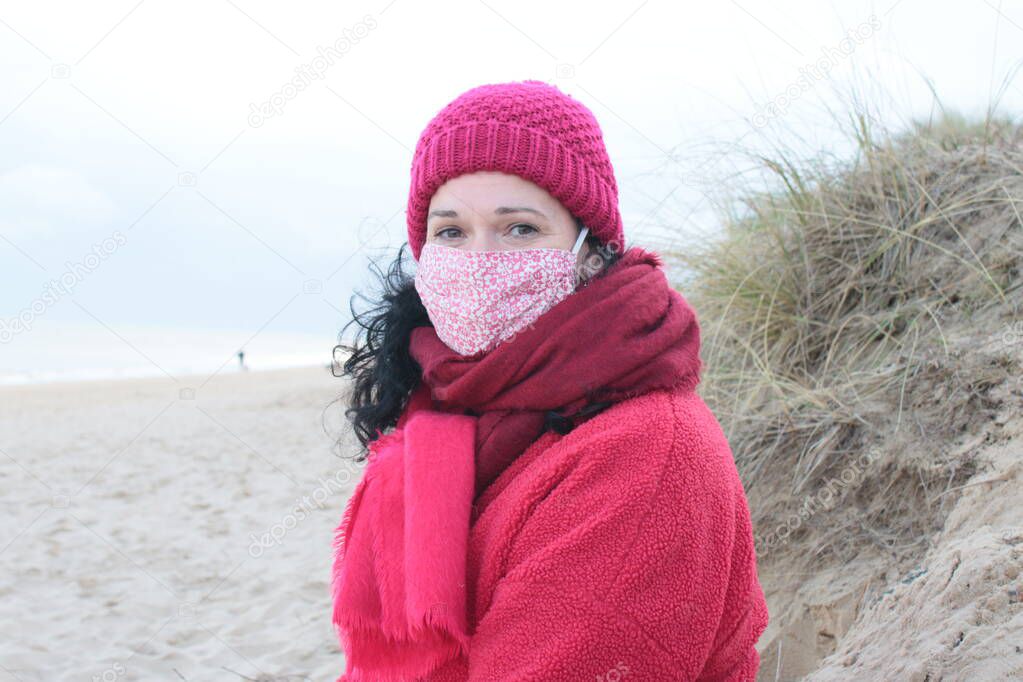 Close up of female wearing mask with black hair in red clothes of red jacket scarf and hat seated on seaside beautiful sandy beach by ocean in Winterton Norfolk east Anglia UK  in lockdown for Covid-19 virus outbreak sea background
