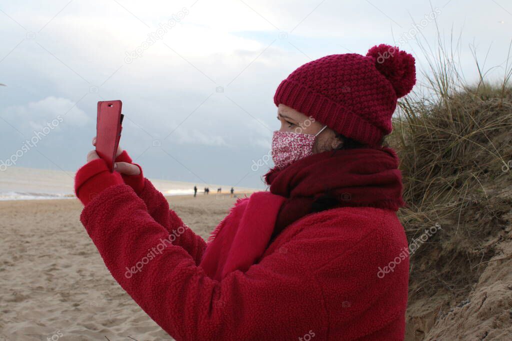 Close up of female wearing mask holding mobile phone  with black hair in red clothes of red jacket scarf and hat seated on seaside beautiful sandy beach by ocean in Winterton Norfolk east Anglia UK  in lockdown Covid-19 virus outbreak sea background