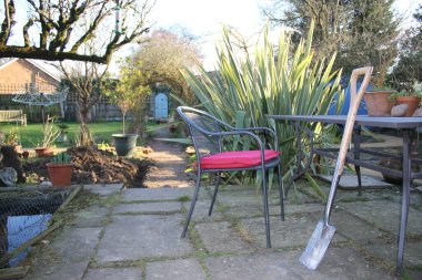 Bahçenin manzarası, masaya yaslanmış yeni bir kürek, verandada kırmızı minder, arka planda gül kemeri, egzotik bitkiler espalier ağacı ve çakıl yolunun sonundaki gizli kapı dikey görüş açısı. 