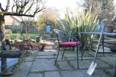 Bahçenin manzarası, masaya yaslanmış yeni bir kürek, verandada kırmızı minder, arka planda gül kemeri, egzotik bitkiler espalier ağacı ve çakıl yolunun sonundaki gizli kapı dikey görüş açısı. 