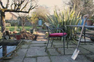 Bahçenin manzarası, masaya yaslanmış yeni bir kürek, verandada kırmızı minder, arka planda gül kemeri, egzotik bitkiler espalier ağacı ve çakıl yolunun sonundaki gizli kapı dikey görüş açısı. 