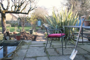 Bahçenin manzarası, masaya yaslanmış yeni bir kürek, verandada kırmızı minder, arka planda gül kemeri, egzotik bitkiler espalier ağacı ve çakıl yolunun sonundaki gizli kapı dikey görüş açısı. 