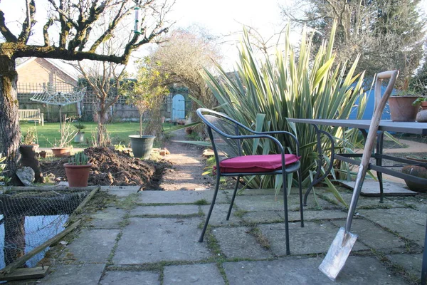 Bahçenin manzarası, masaya yaslanmış yeni bir kürek, verandada kırmızı minder, arka planda gül kemeri, egzotik bitkiler espalier ağacı ve çakıl yolunun sonundaki gizli kapı dikey görüş açısı. 