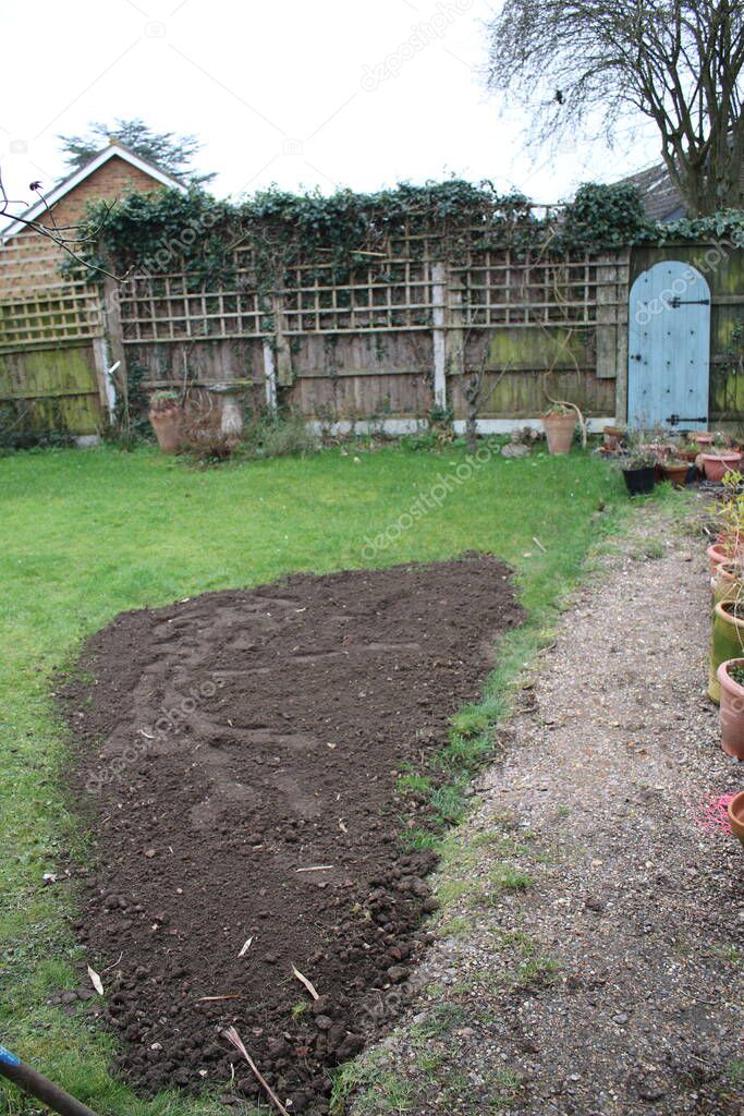 Landscape of organic garden with grass lawn partly covered with fresh soil for re seeding cover, blue door at back fencing with trellis trees, plants and clay plant pots along gravel path in Spring 