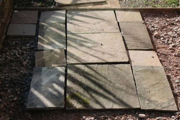Close up of patio construction the sand stone slabs in straight lines on hard core cement and gravel during build with wood edging on the garden path and plants reflected on bricks on warm Spring day