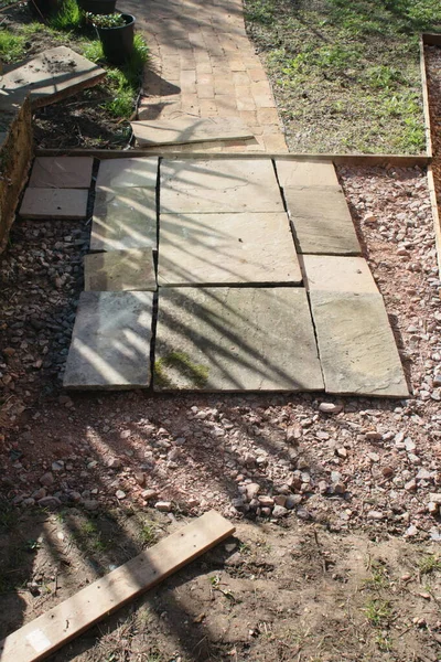 Close up of patio construction the sand stone slabs in straight lines on hard core cement and gravel during build with wood edging on the garden path and plants reflected on bricks on warm Spring day