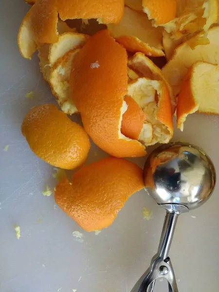 Close up of orange peel, flat lay view the freshly peeled skin of the citrus fruit with yellow pith being prepared to make vegan sweet dessert with a metal ice cream scoop utensil on white kitchen cutting surface