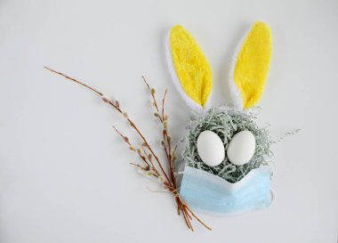 Easter bunny rabbit with yellow ears in a medical mask, branchs pussy willows on a white background with copy space. Creative concept of Easter children's crafts.