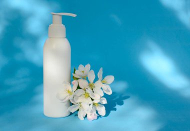 Beyaz kozmetik şişesi ve bahar çiçeklerini gölgeli mavi arka planda taklit et, kopyala alanı. Yüz kavramı, doğal bitki, kozmetik kremi, losyon, şampuan, jöle, sabun, yağ..