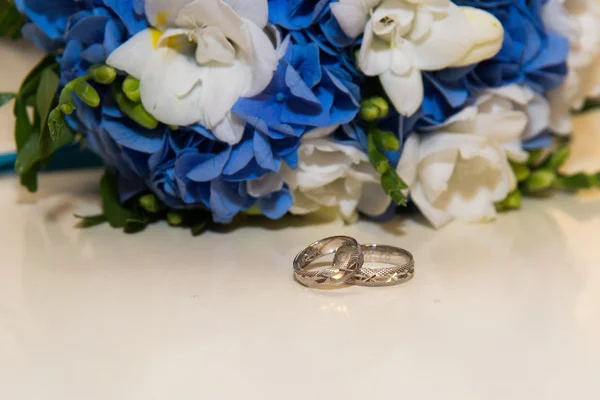 Dois anéis de casamento de platina jazem em torno do buquê da noiva de flores azuis e brancas . — Fotografia de Stock
