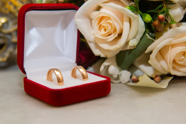 Dois anéis de ouro do casamento com diamantes estão na caixa vermelha perto do buquê da noiva de rosas vermelhas e brancas . — Fotografia de Stock