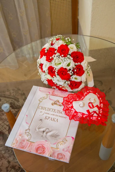 Twee Gouden trouwringen liggen op een kussen in de vorm van een hart met een rode kant rond rode en witte rozen boeket van de bruid. Het certificaat van het huwelijk met een patroon van zwanen en ringen. — Stockfoto
