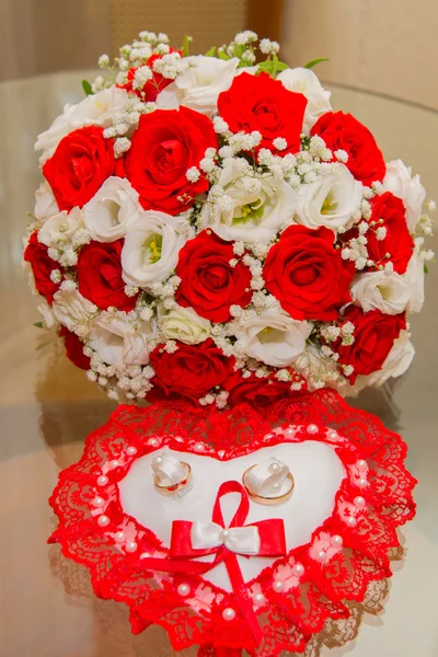 Twee Gouden trouwringen liggen op een kussen in de vorm van een hart met een rode kant rond rode en witte rozen boeket van de bruid. — Stockfoto