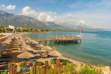 Türkiye 'nin Akdeniz kıyısındaki popüler turizm beldesi Kemer' de güneşli bir günde rıhtımdaki cennet manzarası, şemsiyeler ve güneş dinlenme yerleri ve dağlar.