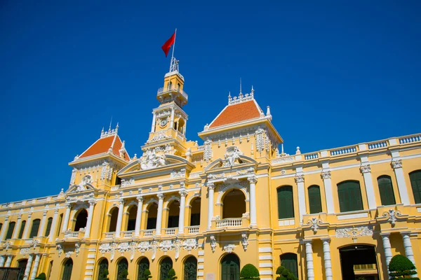 French style of building in Vietnam, Asia. Beautiful Ho Chi Minh City Hall. Facade of house with ornate design. Red flag contrasts with blue sky and clouds. Tourist attraction, famous landmark.