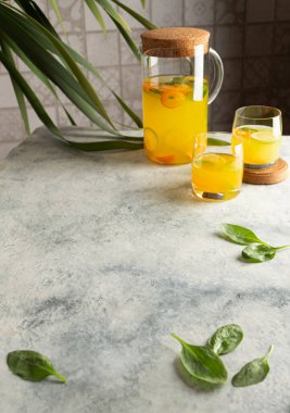 Gri arkaplana uzayı kopyala. Limonatalı gri bir masa, cam sürahide narenciye suyu. Ispanak yaprakları, limon ve limonata dilimleri. Fayans duvarı ve arka planda tropikal bir bitki.