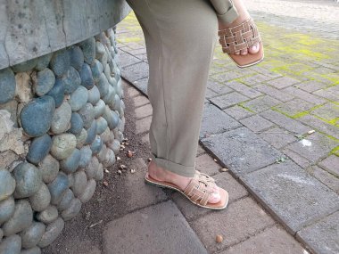 Sarılmış haki pantolonlu, bej örülmüş sandalet giyen, kaldırımda duran, yuvarlak taştan bir duvarın yanında duran insanların alt bacaklarını kapatın.