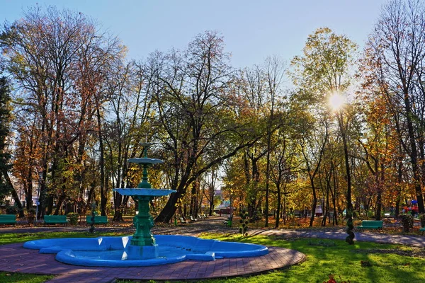 Sonbahar pastel renklerinde bankları olan güzel bir halk parkı çeşmesi ve sokak. Yüksek dinamik aralık fotoğrafçılığı. Dışarıda güneşli bir gün. Güneş ışınları ağaçların arasından parlıyor. Romanya 'nın Gorj ilçesindeki Targu Jiu şehir merkezi turizm merkezi.