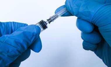 Aşılama. Doktorun mavi eldivenli elleri şırıngayı tutuyor. Şırıngayı aşıya hazırlar. Yakın plan. Beyaz arkaplan.                                