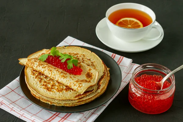 Traditional Russian Crepes Blini stacked in a plate with red caviar and hot lemon tea on wooden background. Maslenitsa traditional Russian festival meal. Russian food, russian kitchen