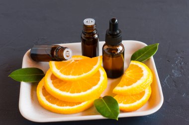 Portakal yağı şişeleri, portakal dilimleri ve yapraklar beyaz tepside ve siyah arka planda. Güzellik, cilt bakımı, refah kavramı