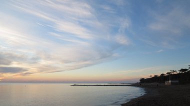 Gökyüzünde ışık bulutları olan Toskana kıyılarının panoramik görüntüsü rüzgar tarafından taşınır, Akdeniz üzerinde günbatımının yoğun renkleri ve kumlu sahil.