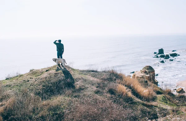 Чоловік стоїть на скелі з собакою-лабрадором і дивиться на море — стокове фото