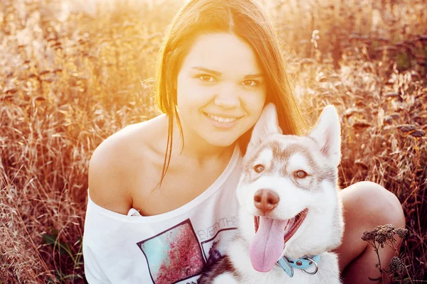 Junge kaukasische Hündin beim Spielen mit ihrem sibirischen Husky-Welpen auf dem Feld während des Sonnenuntergangs. glücklich lächelndes Mädchen, das Spaß mit Welpen im Freien in schönem Licht hat — Stockfoto