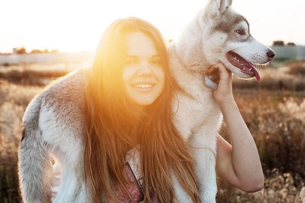 Joven hembra caucásica jugando con su cachorro husky siberiano en el campo durante el atardecer. Feliz chica sonriente divirtiéndose con el cachorro al aire libre en hermosa luz — Foto de Stock