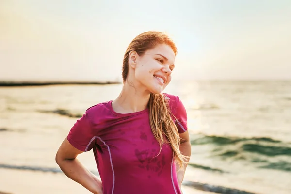 Спортивная модель улыбается радостно делая упражнения на свежем воздухе на восходе солнца. Прекрасные кавказские женские тренировки на берегу моря утром — стоковое фото
