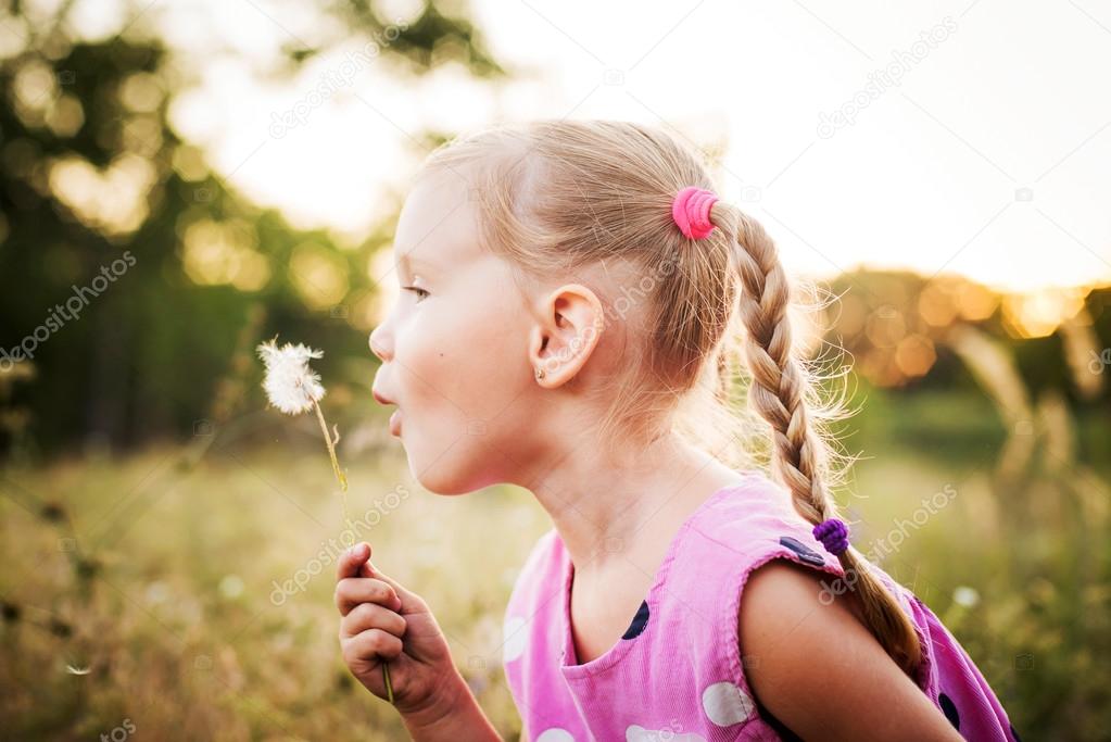 little girl playing in the park in summer day during the sunset, she blowing dandelion on a field
