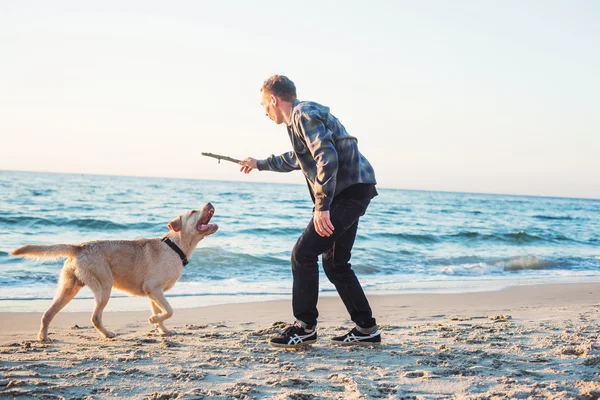 Jovem caucasiano masculino brincando com labrador na praia durante sunri — Fotografia de Stock