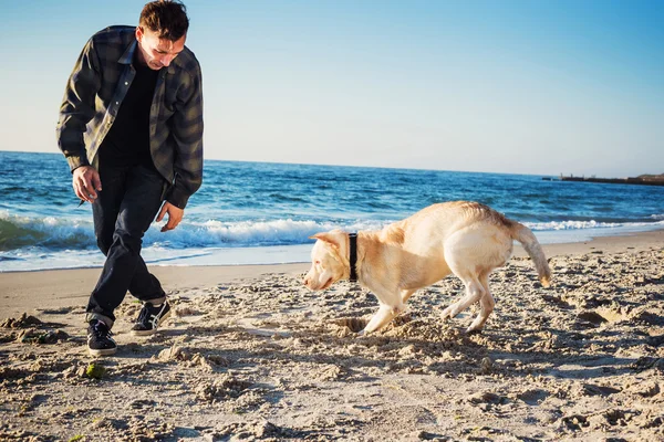 Νεαρά αρσενικά καυκάσιος που παίζει με labrador στην παραλία κατά την Ανατολή το — Φωτογραφία Αρχείου