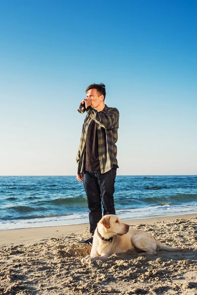 Jovem caucasiano masculino beber café na praia enquanto caminha com — Fotografia de Stock