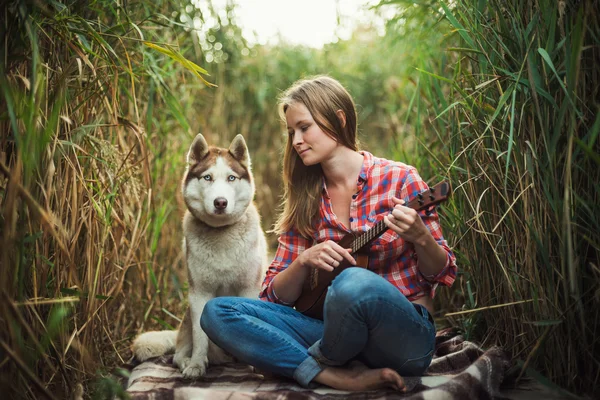 Junge kaukasische Frau spielt Ukulele. Hündin mit sibirischem Husky-Hund spielt im Freien Gitarre — Stockfoto