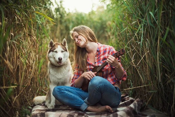 Młoda kobieta kaukaski grając ukulele. Kobieta z psem husky syberyjski, gra na gitarze na zewnątrz — Zdjęcie stockowe
