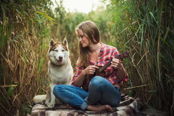 Młoda kobieta kaukaski grając ukulele. Kobieta z psem husky syberyjski, gra na gitarze na zewnątrz — Zdjęcie stockowe