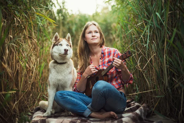 Junge kaukasische Frau spielt Ukulele. Hündin mit sibirischem Husky-Hund spielt im Freien Gitarre — Stockfoto