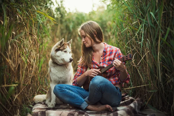 Giovane donna caucasica che suona ukulele. Donna con cane husky siberiano che suona la chitarra all'aperto — Foto Stock