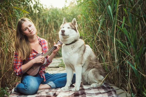 Joven mujer caucásica jugando ukelele. Hembra con perro husky siberiano tocando la guitarra al aire libre — Foto de Stock