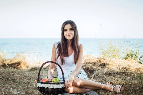 Fêmea caucasiana jovem à beira-mar com cesta com frutas. Menina comendo frutas, alimentação saudável e conceito de vida saudável — Fotografia de Stock