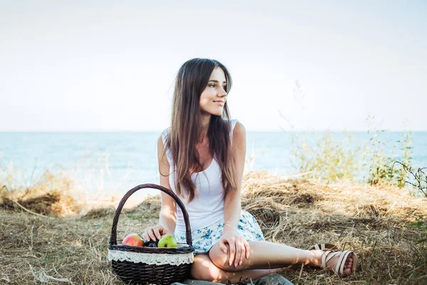 Hembra joven caucásica en la orilla del mar con canasta con frutas. Niña comiendo frutas, alimentación saludable y concepto de vida saludable — Foto de Stock