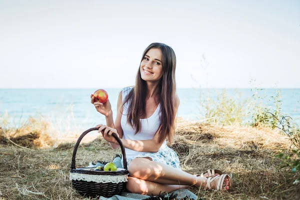 Hembra joven caucásica en la orilla del mar con canasta con frutas. Niña comiendo frutas, alimentación saludable y concepto de vida saludable —  Fotos de Stock