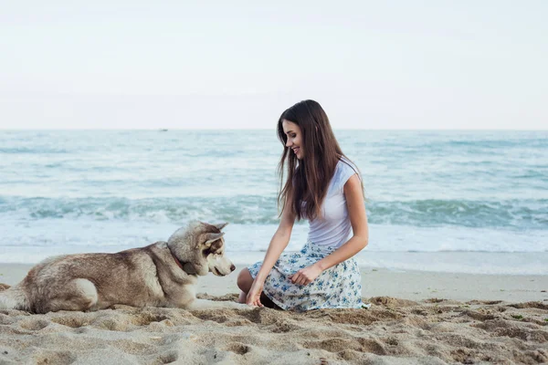 Молодая кавказская женщина играет и веселится с сибирской хаски-собакой на пляже — стоковое фото