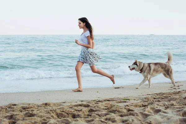Genç beyaz kadın oynamak ve eğlenmek ile Sibirya husky köpek sahilde — Stok fotoğraf