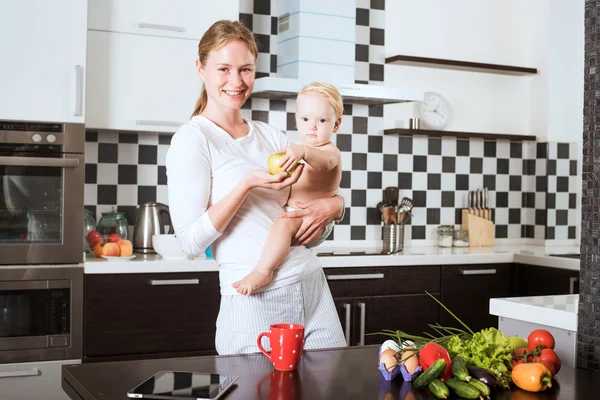 Mother Holding Daughter In Kitchen while cooking a meal of fresh vegetables and fruits. Mother and little daughter on kitchen holding an apple. Healthy food and healthy living concept